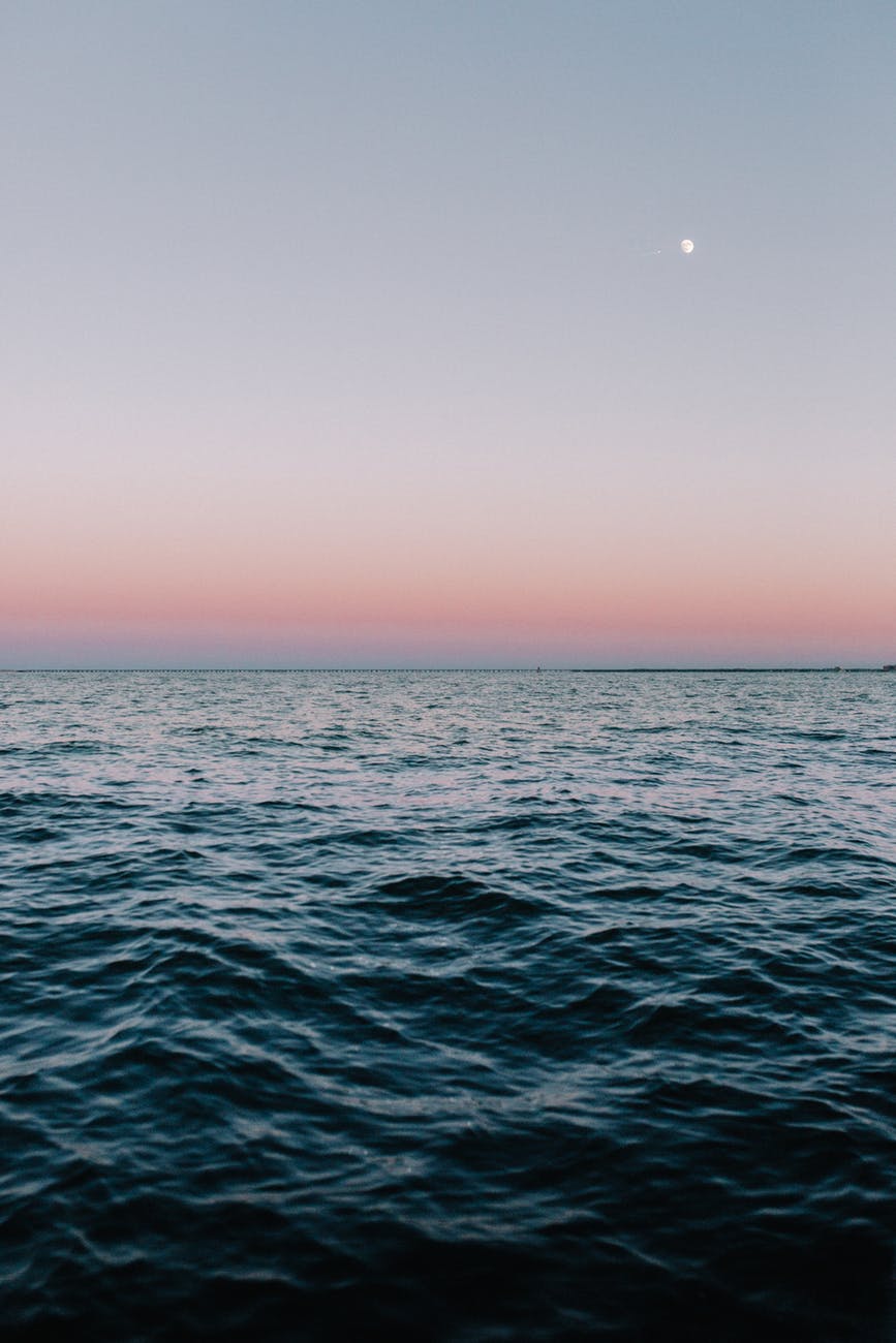 Image Description: A body of water with small waves at dusk. The horizon fades from powdery  blue to powdery pink. A gibbous moon glows in the right corner of the sky.